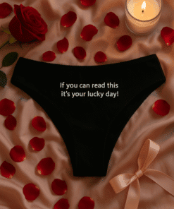Soft black thong with humorous white text, surrounded by rose petals and a lit candle, romantic lingerie gift idea.