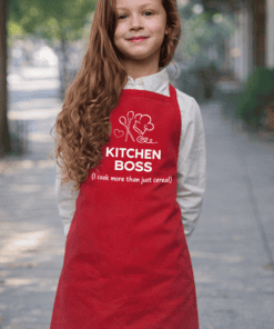 Cute young girl wearing a red kitchen apron with "Kitchen Boss" text and cooking icons, outdoors.