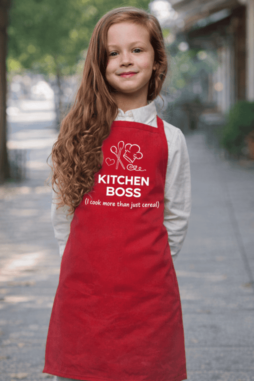 Cute young girl wearing a red kitchen apron with "Kitchen Boss" text and cooking icons, outdoors.