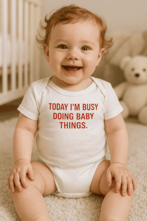 Cute baby wearing a white T-shirt with "Today I'm Busy Doing Baby Things" text, sitting on beige carpet with teddy bear in background.