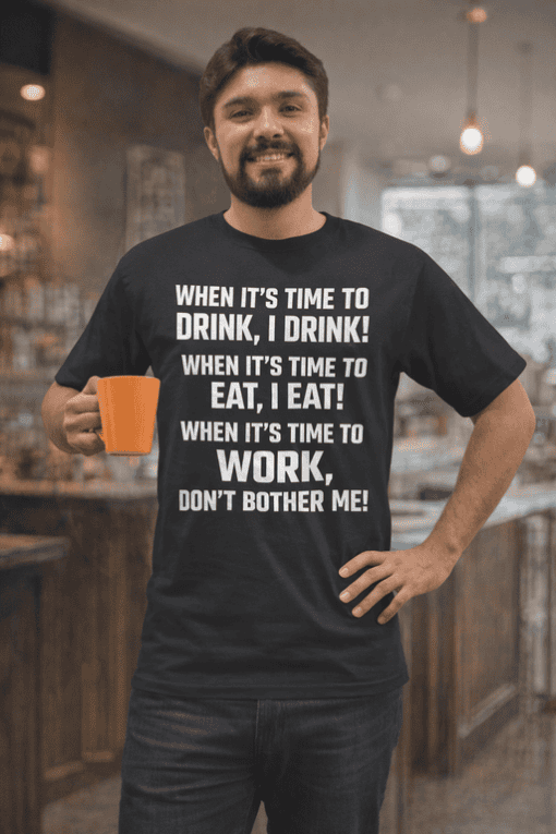 Relaxed man wearing humorous T-shirt about daily routines, holding coffee in a modern cafe setting.