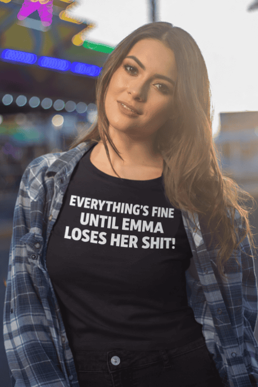 - Woman wearing black T-shirt with humorous text about Emma, casual street style, vibrant nightlife background.
