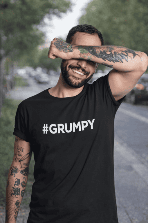 Relaxed man smiling in black "#GRUMPY" T-shirt, casual outdoor setting.