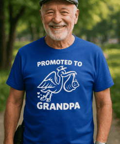 Old man smiling wearing a blue T-shirt with "Promoted to Grandpa" and pelican graphic, outdoors in park setting.