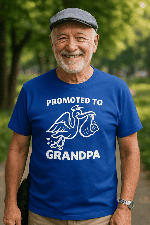Old man smiling wearing a blue T-shirt with "Promoted to Grandpa" and pelican graphic, outdoors in park setting.