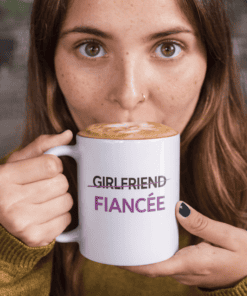 Cute woman holding a coffee mug with "Girlfriend" crossed out and "Fiancée" written, capturing romantic and engagement themes.