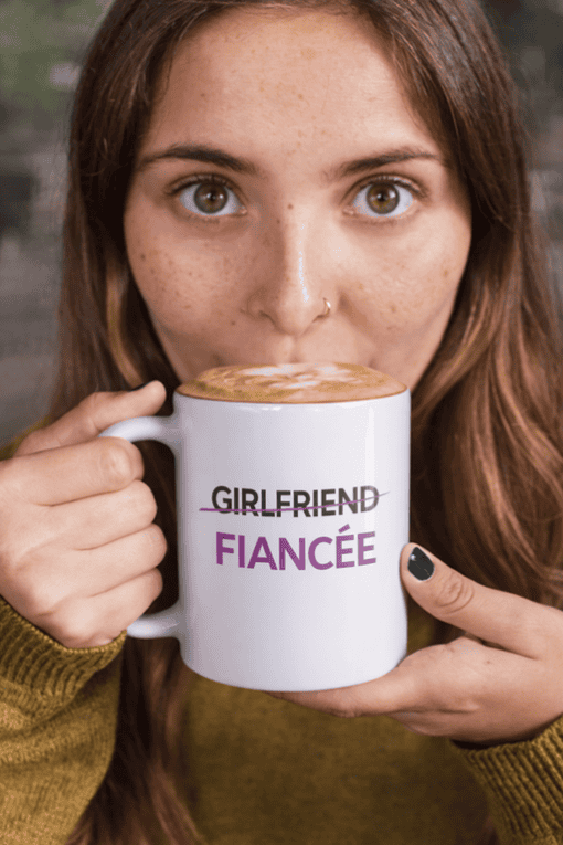 Cute woman holding a coffee mug with "Girlfriend" crossed out and "Fiancée" written, capturing romantic and engagement themes.