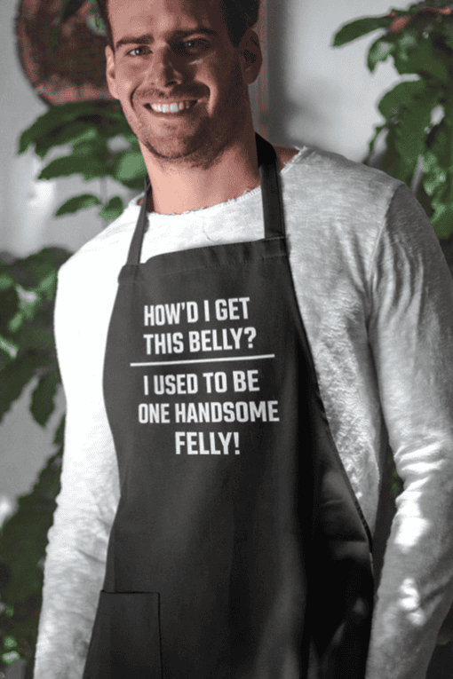 Smiling man wearing humorous apron with text about belly fat in kitchen, casual attire for funny gift idea.