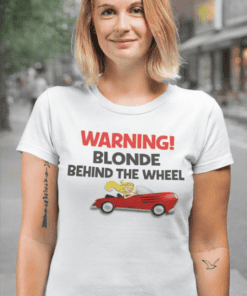 Blonde woman wearing humorous "Warning Blonde Behind the Wheel" T-shirt outdoors.