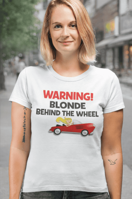 Blonde woman wearing humorous "Warning Blonde Behind the Wheel" T-shirt outdoors.