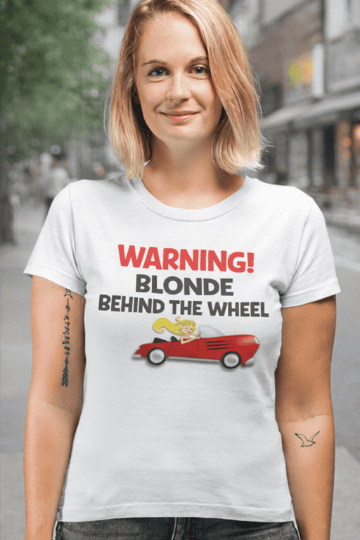 Blonde woman wearing humorous "Warning Blonde Behind the Wheel" T-shirt outdoors.