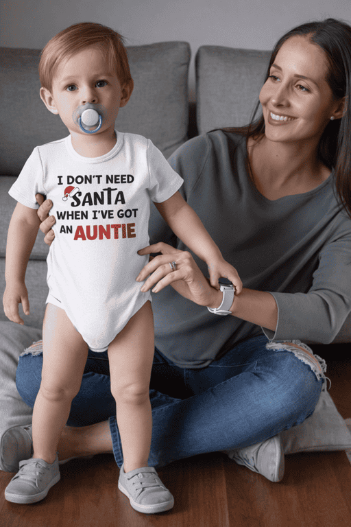 Cute toddler with a pacifier wearing humorous "I DON'T NEED SANTA WHEN I'VE GOT AN AUNTIE" T-shirt for kids, family holiday gift idea.