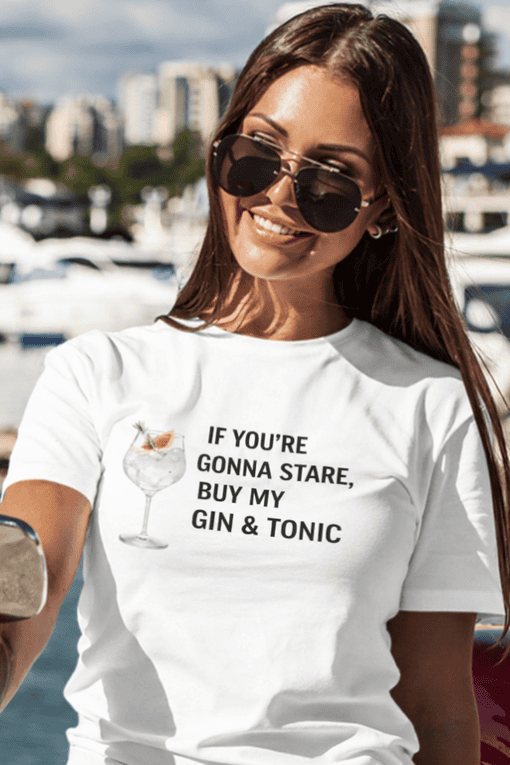 Relaxed woman with sunglasses wearing humorous "Gin & Tonic" slogan T-shirt by the marina, perfect for casual summer fashion.