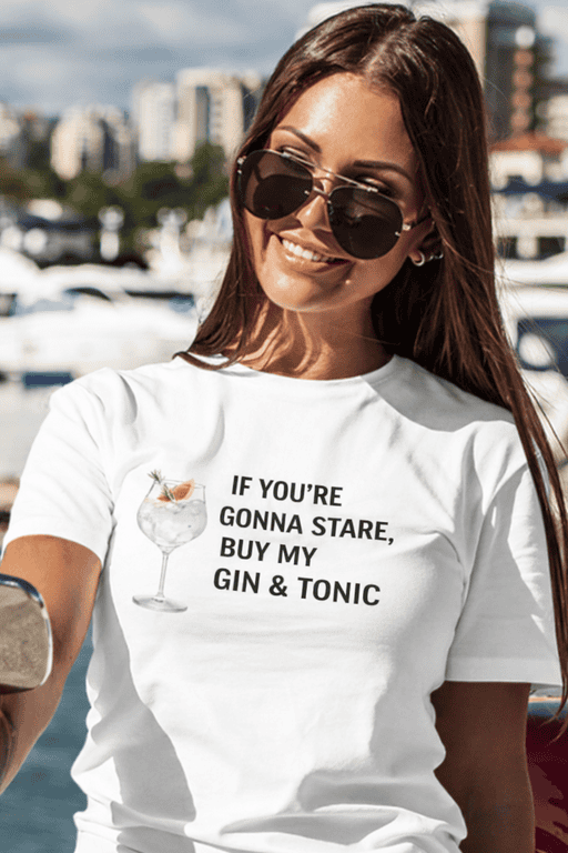 Relaxed woman with sunglasses wearing humorous "Gin & Tonic" slogan T-shirt by the marina, perfect for casual summer fashion.