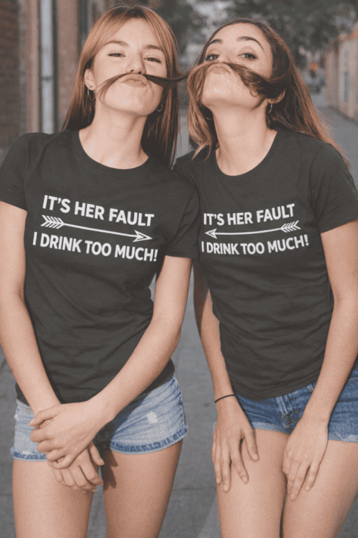 Funny women's T-shirt with "It's her fault I drink too much" phrase, casual quote tee, humorous graphic tee, matching best friend T-shirts, playful matching T-shirt style.
